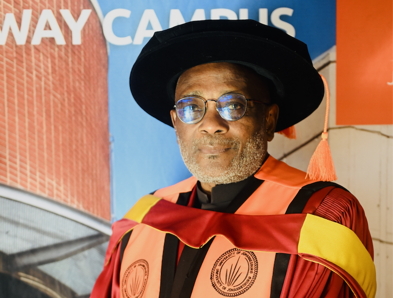 Portrait of a mature man in academic regalia wearing a black graduation cap and orange gown with a yellow sash, posing in front of a campus backdrop.